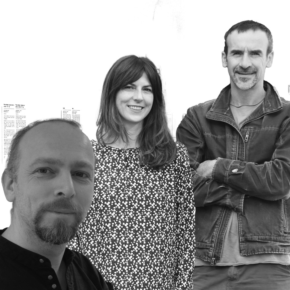 A black and white group portrait of three people, including Olinka Vištica and Dražen Grubišić. In the immediate foreground on the left, a man with short hair and a goatee looks toward the camera. In the center, a woman with long, dark hair and bangs smiles warmly, wearing a dark, patterned blouse. On the right, a man with short hair and a goatee stands with his arms crossed, wearing a dark zippered jacket over a light-colored t-shirt. The background is a bright, nearly white wall with some blurred documents visible.