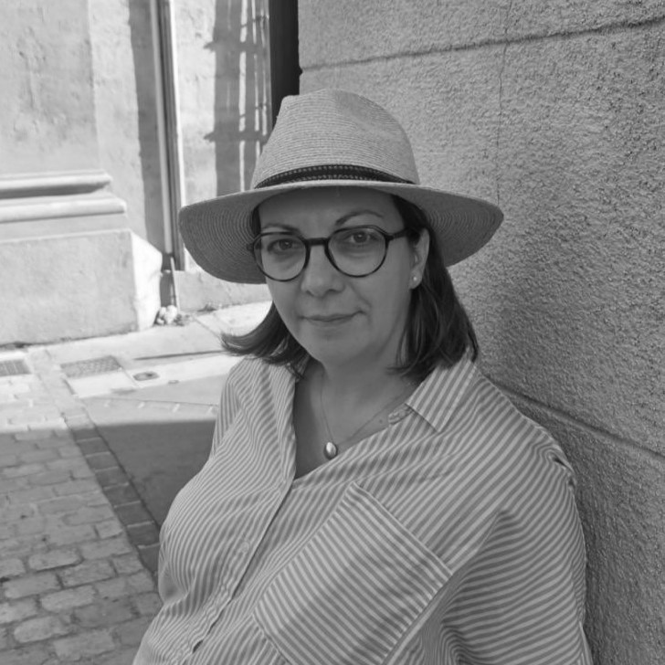 A black and white, medium-shot photograph of a Jovana Diković with dark, shoulder-length hair. She is looking toward the camera with a soft, neutral expression. She is wearing dark-rimmed round glasses, a wide-brimmed hat, and a light-colored button-down shirt with thin vertical stripes. A simple necklace with a small pendant is visible. She is leaning against a textured stone or stucco wall, with a sunlit cobblestone sidewalk and the base of a building visible in the background.